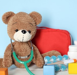 Photo of Pediatrician concept. Toy bear, cubes, first aid kit, pills, thermometer and stethoscope on white wooden table against light blue background, closeup