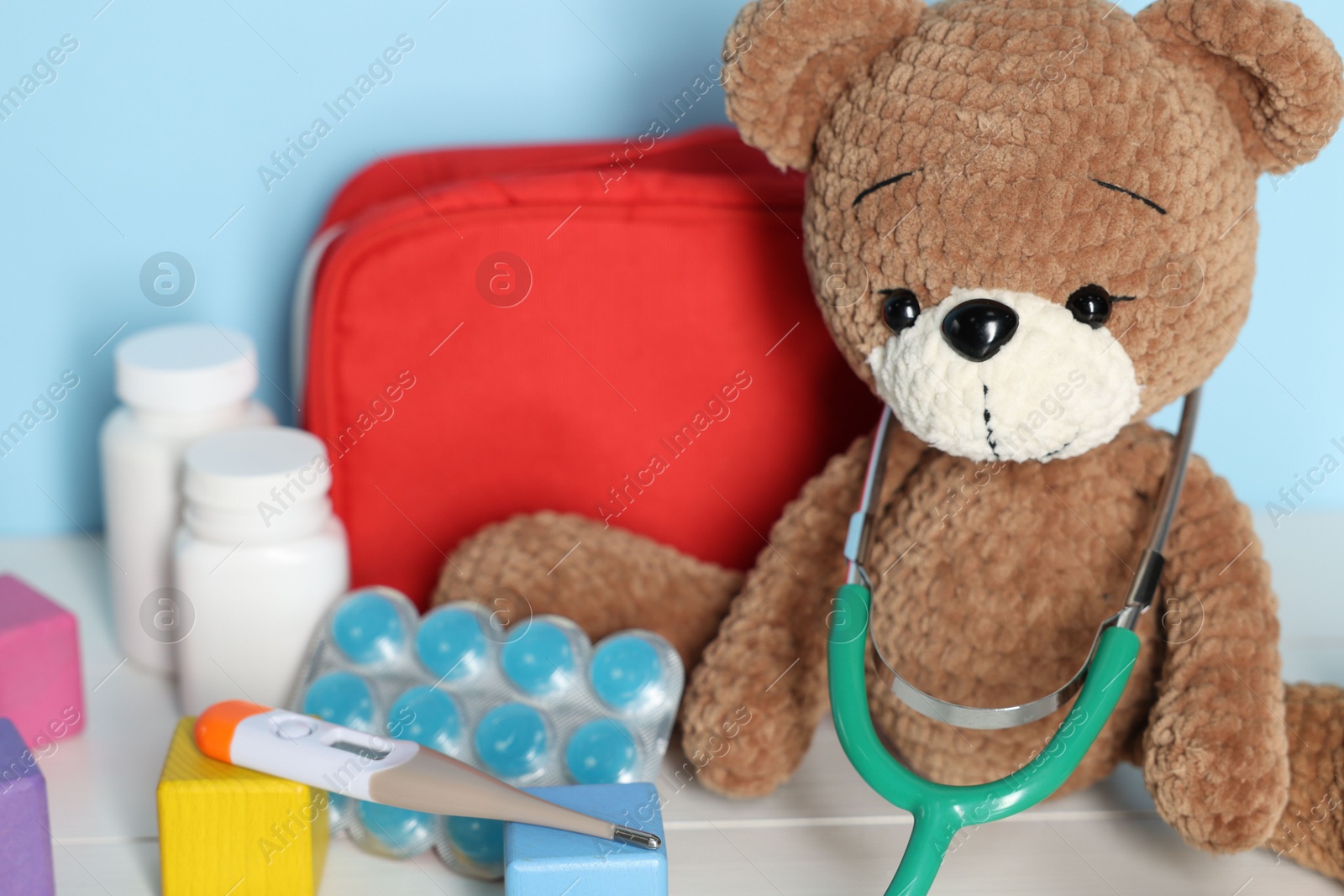 Pediatrician concept. Toy bear, cubes, first aid kit, pills, thermometer and stethoscope on white wooden table against light blue background, closeup Photo of Pediatrician concept. Toy bear, cubes, first aid kit, pills, thermometer and stethoscope on white wooden table against light blue background, closeup