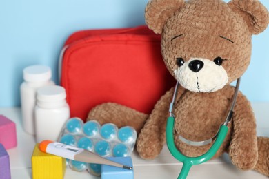 Photo of Pediatrician concept. Toy bear, cubes, first aid kit, pills, thermometer and stethoscope on white wooden table against light blue background, closeup