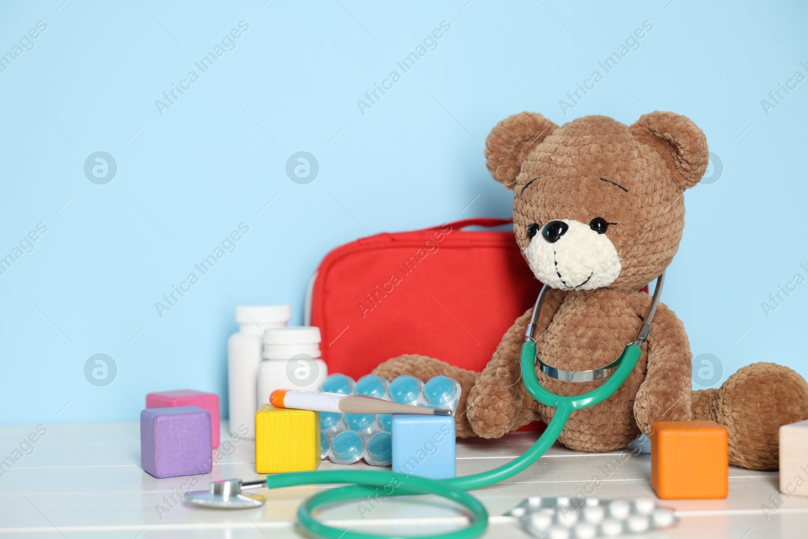Pediatrician concept. Toy bear, cubes, first aid kit, pills and stethoscope on white wooden table against light blue background Photo of Pediatrician concept. Toy bear, cubes, first aid kit, pills and stethoscope on white wooden table against light blue background