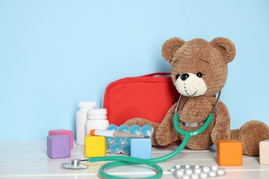 Photo of Pediatrician concept. Toy bear, cubes, first aid kit, pills and stethoscope on white wooden table against light blue background