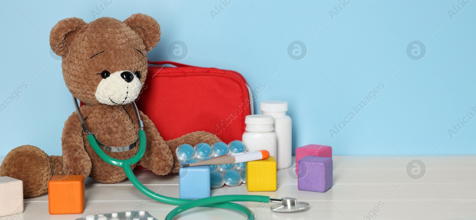 Pediatrician concept. Toy bear, cubes, first aid kit, pills and stethoscope on wooden table against light blue background, space for text Photo of Pediatrician concept. Toy bear, cubes, first aid kit, pills and stethoscope on wooden table against light blue background, space for text