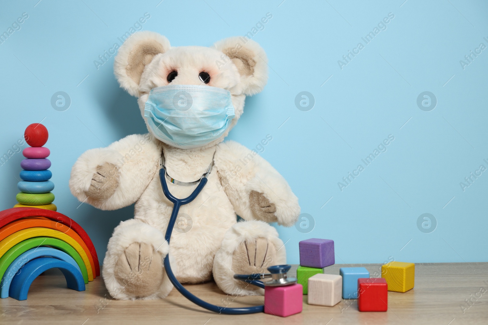 Pediatrician concept. Toys, medical mask and stethoscope on wooden table against light blue background, space for text Photo of Pediatrician concept. Toys, medical mask and stethoscope on wooden table against light blue background, space for text