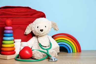Photo of Pediatrician concept. Toys, stethoscope, first aid kit, red heart and pills on wooden table against light blue background