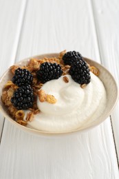 Photo of Tasty yogurt with granola and blackberries in bowl on white wooden table, closeup