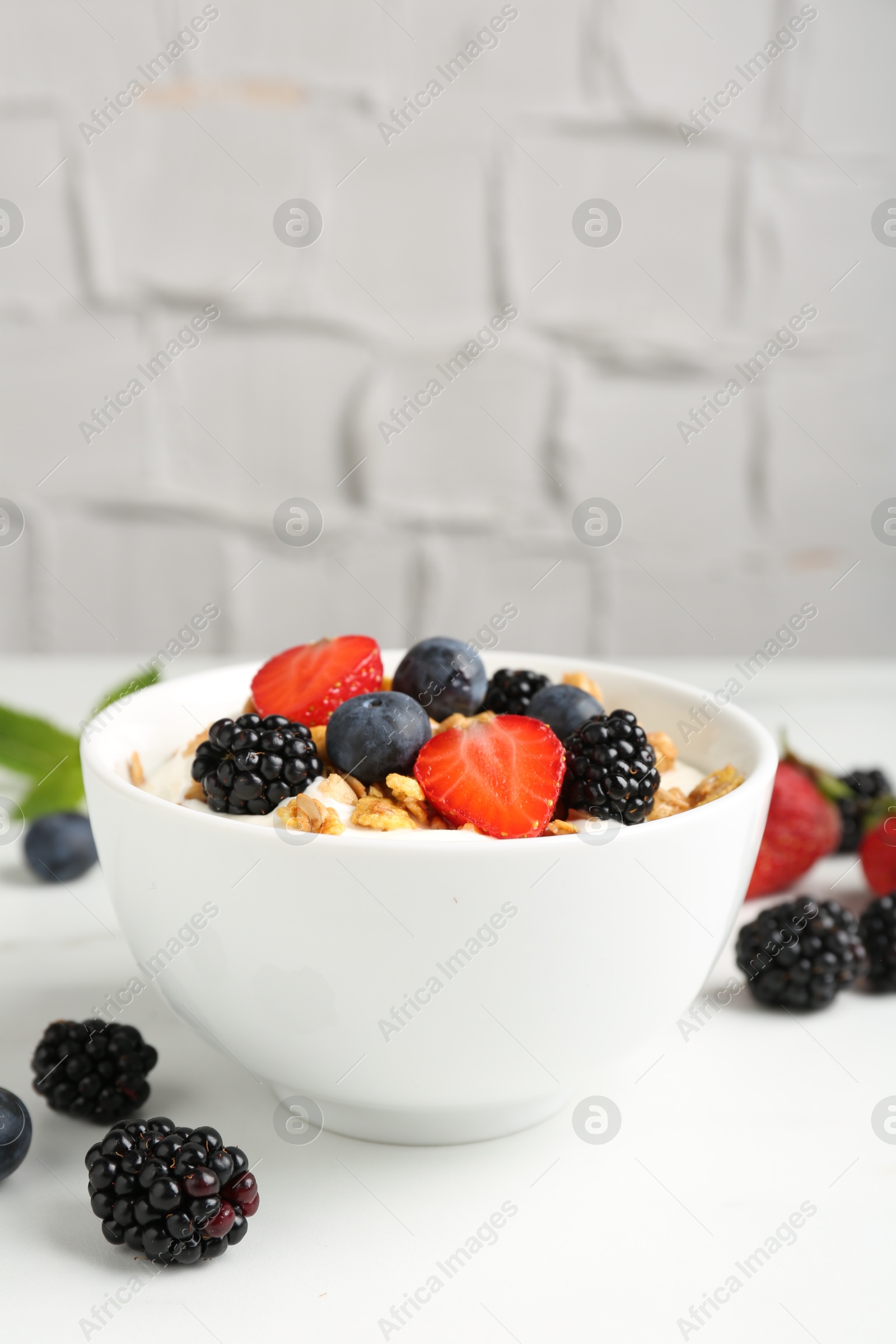 Photo of Tasty yogurt with granola and berries in bowl on white table, closeup. Space for text
