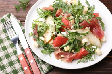 Photo of Tasty salad with arugula, prosciutto, cheese, pear, grapefruit and nuts on wooden table, above view