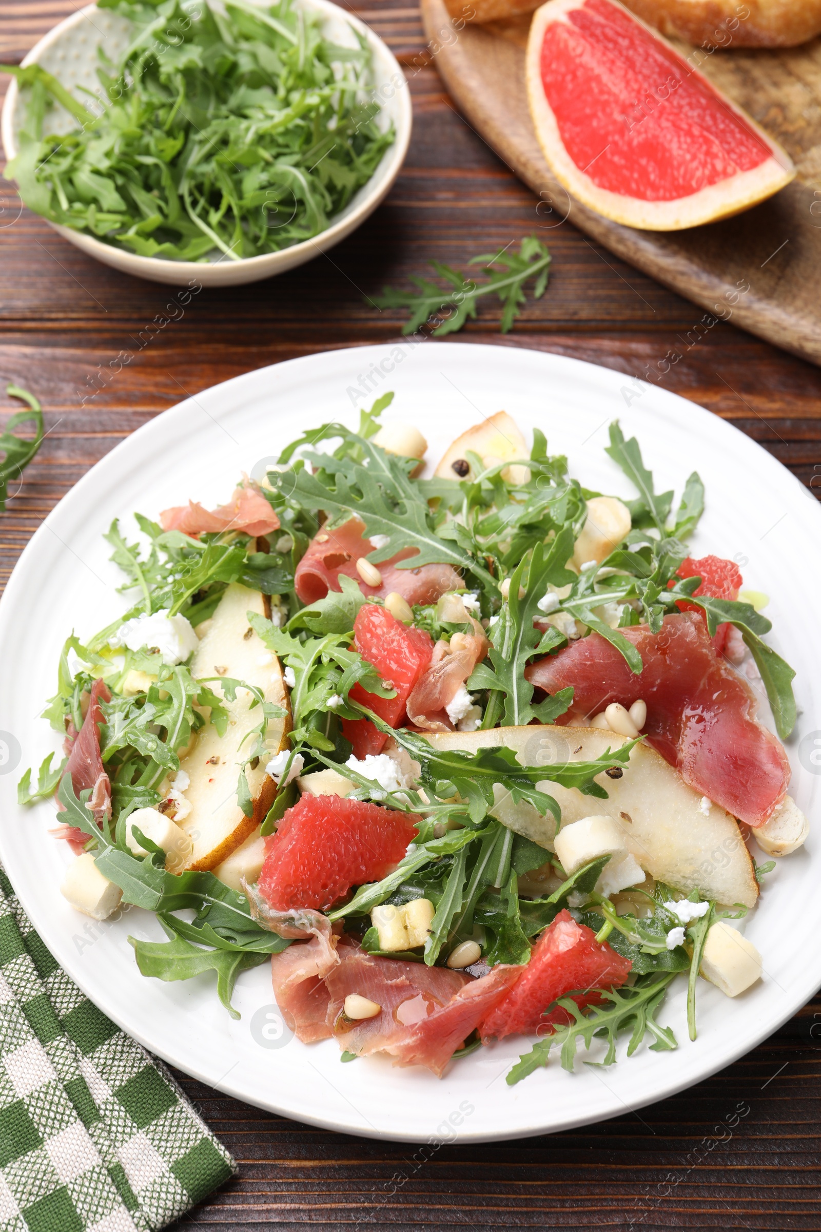 Photo of Tasty salad with arugula, prosciutto, cheese, pear, grapefruit and nuts on wooden table, above view