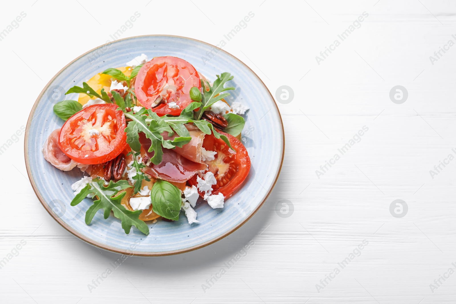Photo of Tasty salad with arugula, cheese, tomatoes, nuts and prosciutto on white wooden table, above view. Space for text