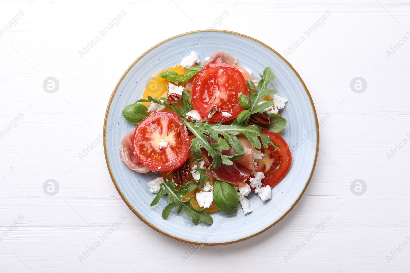 Photo of Tasty salad with arugula, cheese, tomatoes, nuts and prosciutto on white wooden table, top view