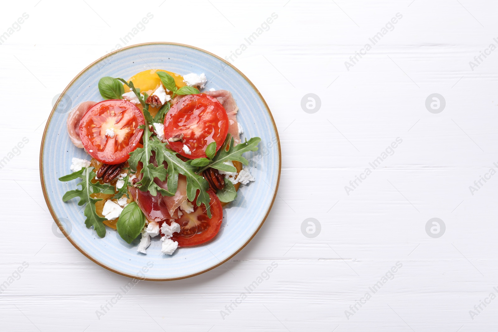 Photo of Tasty salad with arugula, cheese, tomatoes, nuts and prosciutto on white wooden table, top view. Space for text