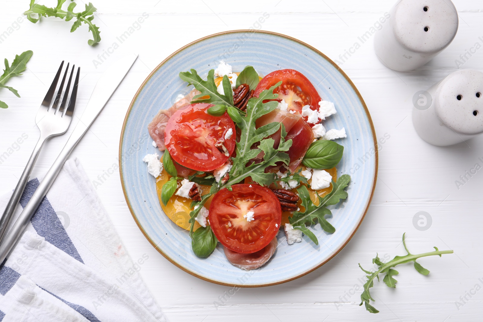 Photo of Tasty salad with arugula, cheese, tomatoes, nuts and prosciutto on white wooden table, flat lay