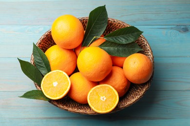 Photo of Fresh oranges and leaves in wicker basket on light blue wooden table, top view