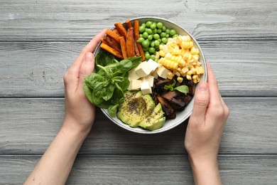 Photo of Healthy vegetarian food. Woman with delicious Buddha bowl at grey wooden table, top view