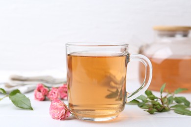 Photo of Aromatic tea in glass cup, roses and green leaves on white table, closeup