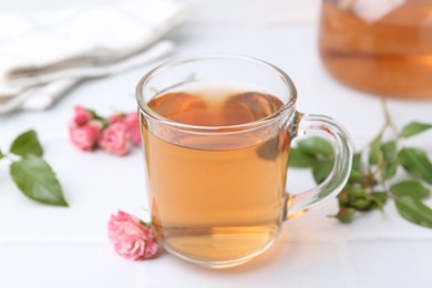 Photo of Aromatic tea in glass cup, roses and green leaves on white table, closeup