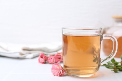 Photo of Aromatic tea in glass cup, roses and green leaves on white table, closeup. Space for text