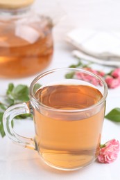 Photo of Aromatic tea in glass cup, roses and green leaves on white table, closeup