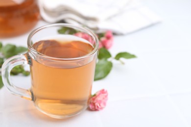 Photo of Aromatic tea in glass cup, roses and green leaves on white table, closeup. Space for text
