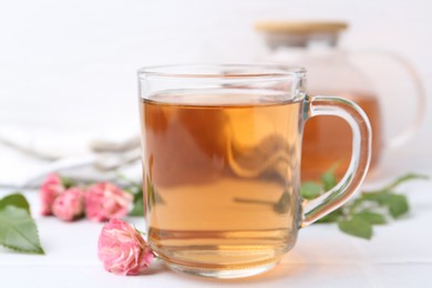 Photo of Aromatic tea in glass cup, roses and green leaves on white table, closeup