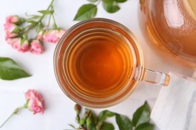 Photo of Aromatic tea in glass cup, roses and green leaves on white table, flat lay