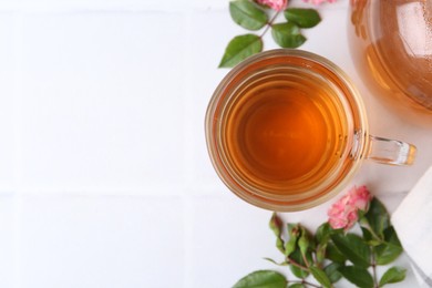 Photo of Aromatic tea in glass cup, roses and green leaves on white table, flat lay. Space for text