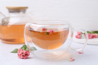Photo of Aromatic tea in glass cup, teapot, roses and green leaves on light table, closeup