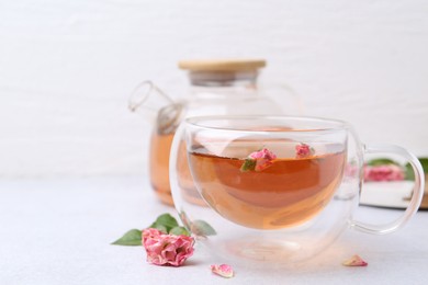 Photo of Aromatic tea in glass cup, teapot, roses and green leaves on light table, closeup. Space for text