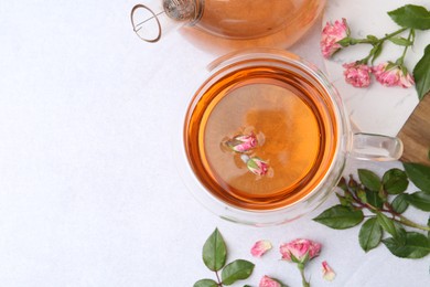 Photo of Aromatic tea in glass cup, teapot, roses and green leaves on light table, flat lay. Space for text