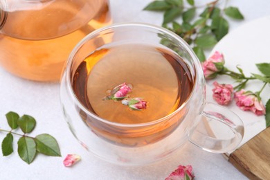 Photo of Aromatic tea in glass cup, teapot, roses and green leaves on light table, closeup