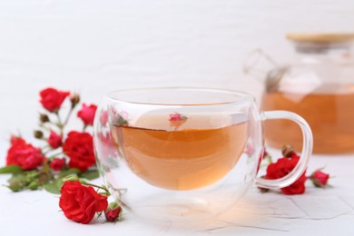 Photo of Aromatic tea in glass cup, roses and green leaves on white table, closeup