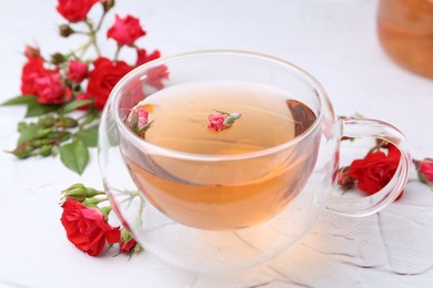 Photo of Aromatic tea in glass cup, roses and green leaves on white table, closeup