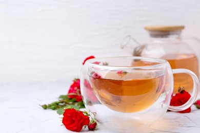 Photo of Aromatic tea in glass cup, roses and green leaves on white table, closeup. Space for text