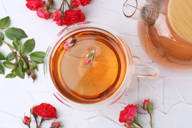 Photo of Aromatic tea in glass cup, teapot, roses and green leaves on white table, flat lay