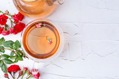 Photo of Aromatic tea in glass cup, teapot, roses and green leaves on white table, flat lay. Space for text