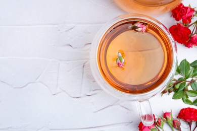 Photo of Aromatic tea in glass cup, roses and green leaves on white table, flat lay. Space for text