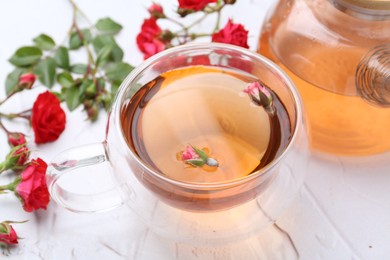 Photo of Aromatic tea in glass cup, teapot, roses and green leaves on white table, closeup