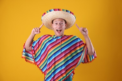 Photo of Emotional man in colorful poncho pointing at his sombrero hat on yellow background
