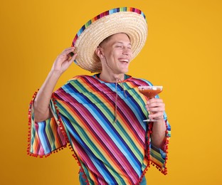 Photo of Happy man in sombrero hat, colorful poncho and cocktail on yellow background