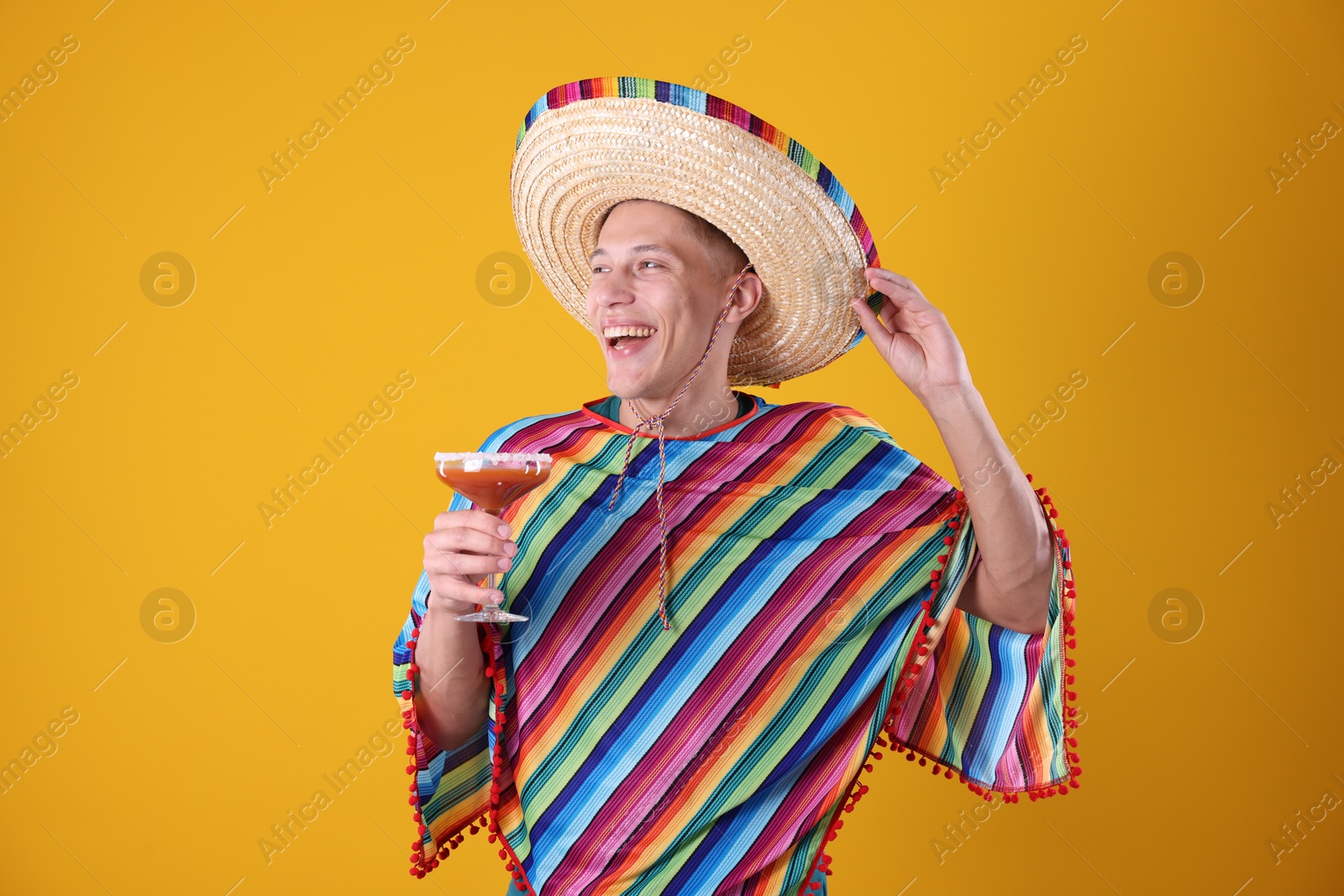 Photo of Happy man in sombrero hat, colorful poncho and cocktail on yellow background
