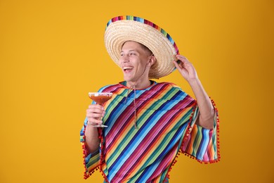 Photo of Happy man in sombrero hat, colorful poncho and cocktail on yellow background