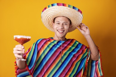 Photo of Happy man in sombrero hat, colorful poncho and cocktail on yellow background