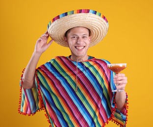 Photo of Happy man in sombrero hat, colorful poncho and cocktail on yellow background