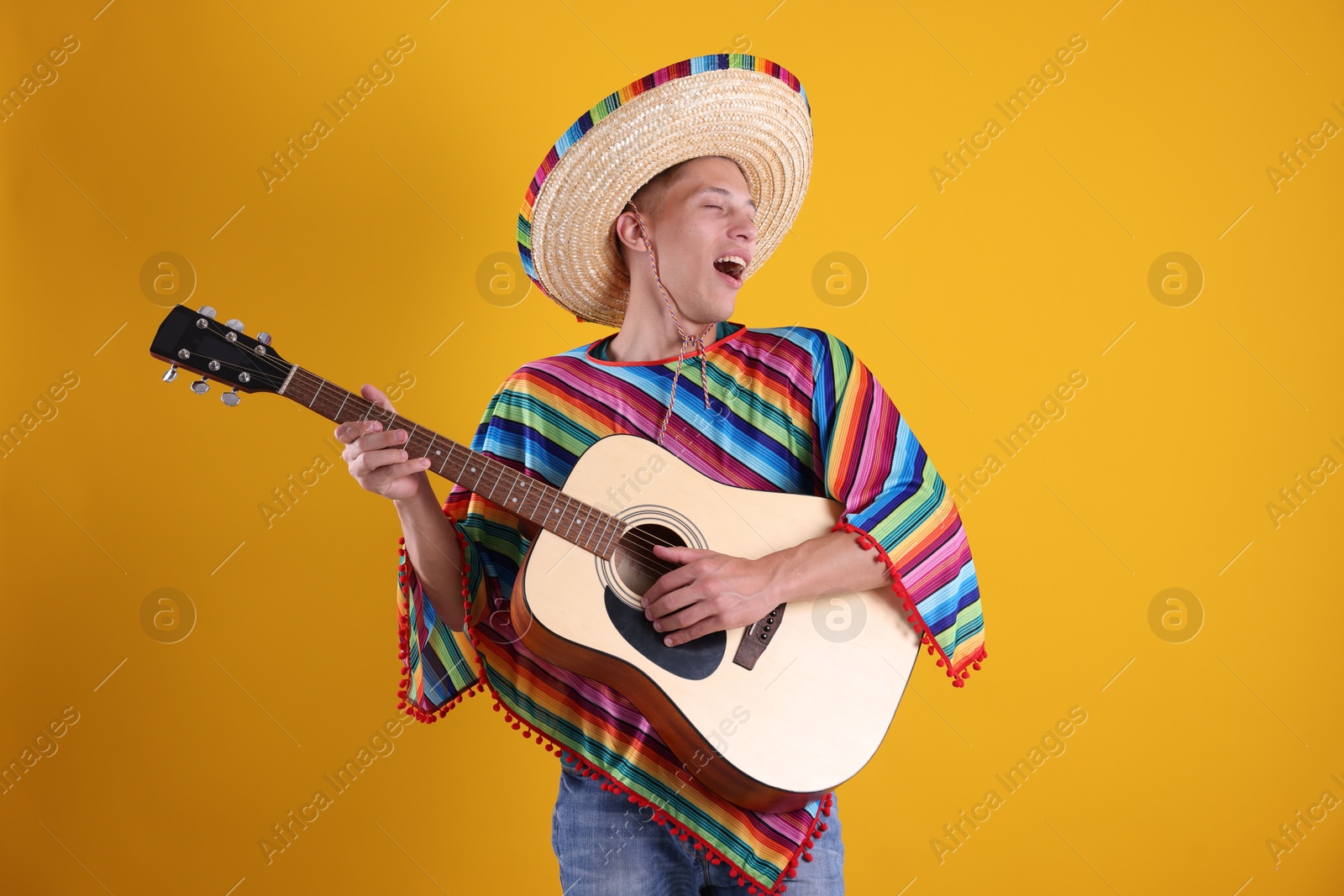 Photo of Happy man in sombrero hat and colorful poncho playing guitar on yellow background