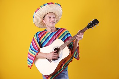 Photo of Happy man in sombrero hat and colorful poncho playing guitar on yellow background