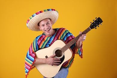 Photo of Happy man in sombrero hat and colorful poncho playing guitar on yellow background