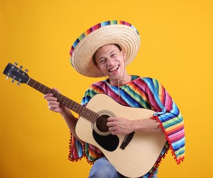 Photo of Happy man in sombrero hat and colorful poncho playing guitar on yellow background