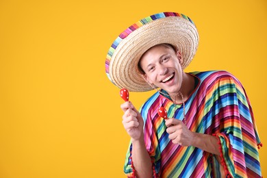 Photo of Happy man in sombrero hat and colorful poncho playing maracas on yellow background. Space for text