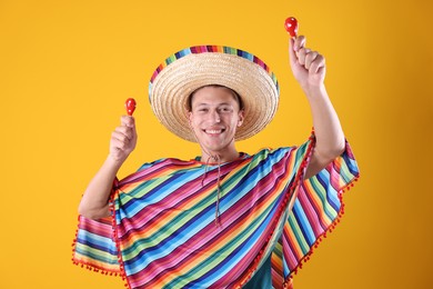 Photo of Happy man in sombrero hat and colorful poncho playing maracas on yellow background