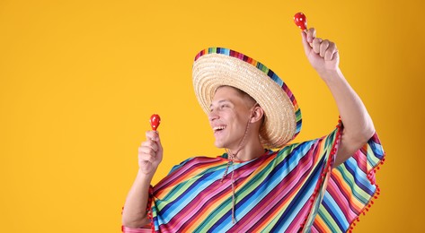 Photo of Happy man in sombrero hat and colorful poncho playing maracas on yellow background. Space for text